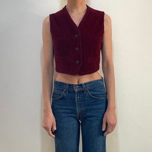 Può includere: Un gilet di velluto a coste bordeaux corto con chiusura a bottoni e due tasche. Il gilet è indossato sopra un paio di jeans blu.