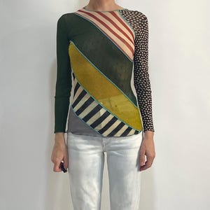 Può includere: Un top a maniche lunghe con un design patchwork composto da verde scuro a tinta unita, strisce diagonali rosse e bianche, un pannello testurizzato giallo senape, strisce diagonali bianche e nere e una manica in rete nera con pois bianchi, abbinato a jeans azzurri.