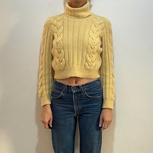 Puede incluir: Un suéter amarillo de punto de cable corto con mangas largas. El suéter se lleva con jeans azules.