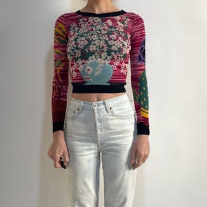 Può includere: Un maglione cropped a maniche lunghe con un design floreale e jeans azzurri. Il maglione presenta un vivace motivo floreale nei toni del rosa, verde e blu. I jeans sono in denim chiaro con gamba dritta.