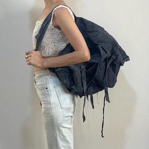May include: A large, dark gray backpack with multiple straps and pockets. The backpack appears to be made of a durable, possibly canvas-like material. The straps are long and adjustable. The backpack is worn over the shoulder.