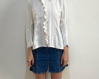 May include: A white long-sleeve blouse with a high collar and ruffled detailing down the front. The blouse features lace inserts and is paired with a denim skirt. The outfit is simple and classic.