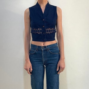 Può includere: Un gilet senza maniche in denim blu scuro con scollo a V e chiusura con bottoni. Il gilet presenta un pannello traforato decorativo sull'addome. La modella indossa anche jeans blu.