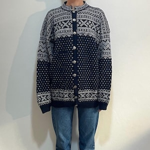 Könnte beinhalten: Eine marineblaue Strickjacke mit weißem geometrischen Muster und silbernen Knöpfen. Die Strickjacke wird mit einer blauen Jeans kombiniert. Der Pullover hat einen hohen Kragen und eine Knopfleiste. Die Jeans ist im klassischen Straight-Leg-Stil.