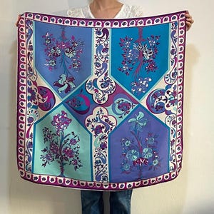 May include: A large square scarf with a floral design in shades of blue, purple, and teal. The scarf features a geometric pattern with stylized flowers and leaves. The border is a deep purple with a repeating design.