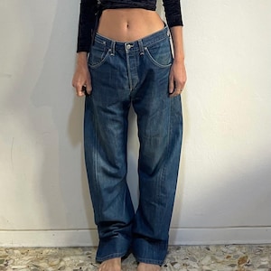 Puede incluir: Un top cruzado de terciopelo negro con mangas largas y cuello en V profundo, combinado con vaqueros de mezclilla azul descolorido de corte holgado. Los vaqueros tienen bragueta con botones y un ajuste relajado. El atuendo es casual y elegante.