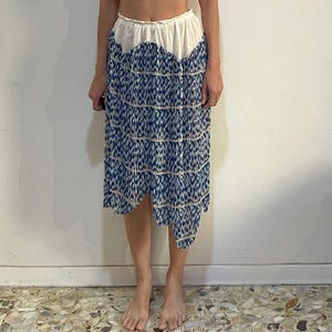 Op de afbeelding: Een witte, mouwloze, knoopsluiting top met kanten details wordt gecombineerd met een blauw-wit patroon rok. De rok heeft een unieke asymmetrische zoom. De outfit is een vintage stijl.