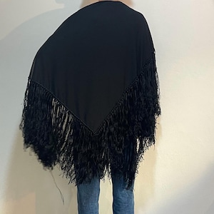 Puede incluir: Un chal negro con flecos largos. El chal está drapeado sobre los hombros y tiene forma triangular. Los flecos cuelgan de los bordes del chal. La persona lleva vaqueros azules.