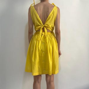 May include: A bright yellow sleeveless dress featuring a deep V-neckline with tie-shoulder straps, an open back with a central tie closure, and a gathered skirt with a vertical row of buttons running down the center of the back panel.