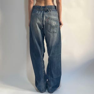 Puede incluir: Un top corto de canalé verde teñido anudado y unos vaqueros de mezclilla azul descolorido. El top es sin mangas y los vaqueros tienen un corte de pierna ancha. Los vaqueros tienen una etiqueta de Levi's en el bolsillo trasero.