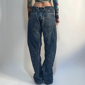 Puede incluir: Una persona que lleva un top corto de manga larga teñido anudado en tonos azules, verdes, amarillos y rojos, combinado con vaqueros de mezclilla de lavado oscuro. Los vaqueros tienen un diseño único con cintura doblada y corte holgado.