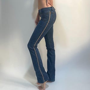 May include: Blue denim jeans with a flared leg and a decorative gold-colored trim along the side seams. The jeans are paired with a white, sleeveless, lace-trimmed top. The outfit is displayed against a plain white background.