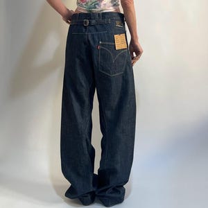 Puede incluir: Vaqueros de mezclilla azul oscuro con pernera ancha y cintura alta. Los vaqueros tienen un parche de cuero y una pequeña etiqueta roja en el bolsillo trasero. La modelo lleva un top de tirantes con estampado floral. Los vaqueros tienen cinturón y hebilla.