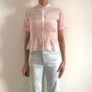 Pu&ograve; includere: Una blusa rosa a maniche corte con ricami bianchi sul colletto e sul carr&eacute;. La blusa ha una chiusura con bottoni sul davanti e una vita aderente.