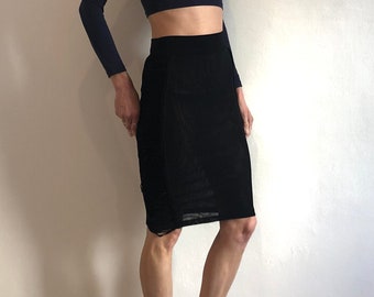 May include: A woman is wearing a navy blue long-sleeved crop top and a black skirt. She is standing with her feet slightly apart and her arms at her sides.