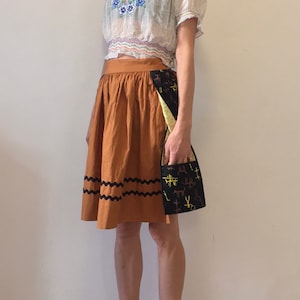 Puede incluir: Una mujer lleva una blusa blanca bordada con una falda marrón con ribete negro. Lleva un bolso estampado en negro y amarillo.