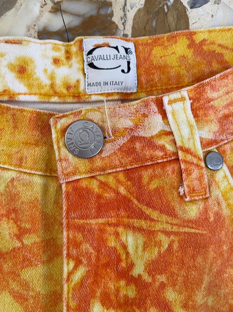 May include: Orange and yellow tie-dye denim jeans with a Cavalli Jeans label and a metal button with the Cavalli Jeans logo. The label reads "Cavalli Jeans" and "Made in Italy".