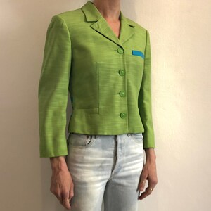 Pode incluir: Um blazer verde curto com um detalhe de acabamento azul. O blazer tem um fecho de bot&otilde;es e um &uacute;nico bolso no lado esquerdo.