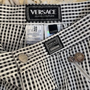 May include: Close-up of Versace Jeans Couture pants. The black and white checkered pants feature a zipper, a button, and multiple tags with the Versace logo. The pants are made of a blend of polyamide and spandex.