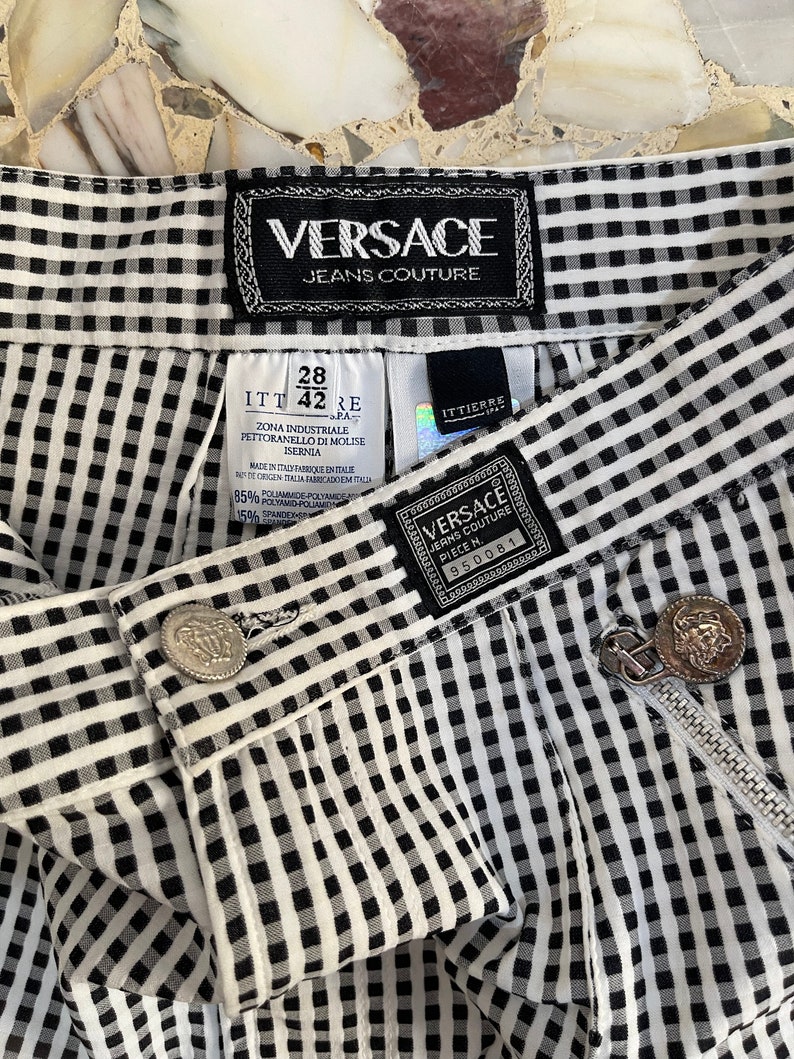 May include: Close-up of Versace Jeans Couture pants. The black and white checkered pants feature a zipper, a button, and multiple tags with the Versace logo. The pants are made of a blend of polyamide and spandex.