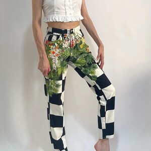 May include: High-waisted denim jeans featuring a bold black and white checkerboard pattern. The front of the jeans is decorated with a vibrant, detailed print of green palm leaves and colorful tropical flowers layered over the checkered design.