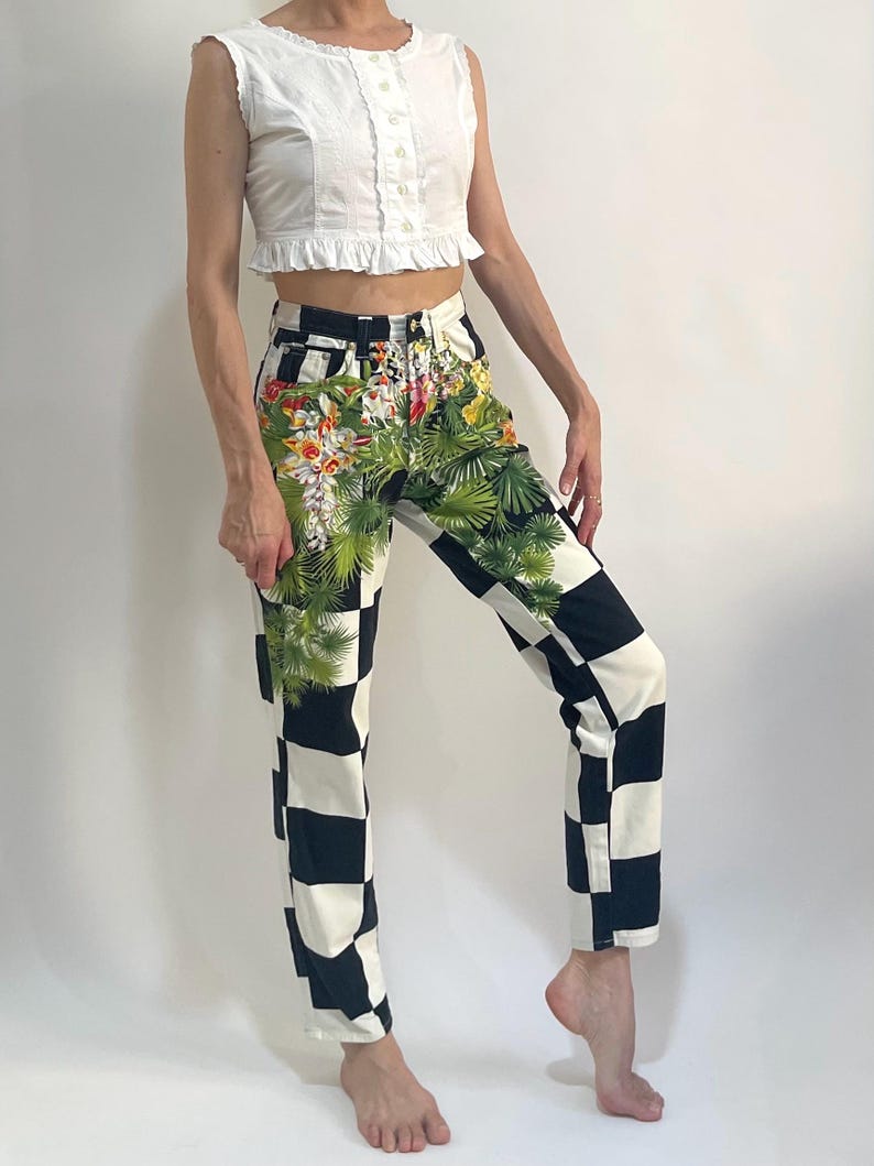 May include: High-waisted denim jeans featuring a bold black and white checkerboard pattern. The front of the jeans is decorated with a vibrant, detailed print of green palm leaves and colorful tropical flowers layered over the checkered design.