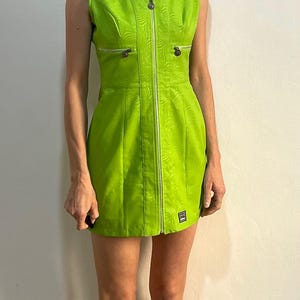 Może przedstawiać: Kr&oacute;tka, limonkowo-zielona sukienka bez rękaw&oacute;w z zamkiem błyskawicznym z przodu i subtelnym tłoczonym wzorem. Sukienka ma dwie kieszenie z przodu z srebrnymi zamkami błyskawicznymi.