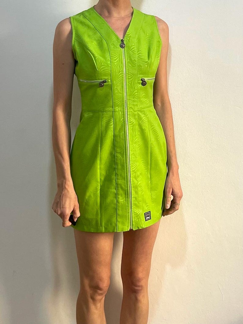 Może przedstawiać: Kr&oacute;tka, limonkowo-zielona sukienka bez rękaw&oacute;w z zamkiem błyskawicznym z przodu i subtelnym tłoczonym wzorem. Sukienka ma dwie kieszenie z przodu z srebrnymi zamkami błyskawicznymi.