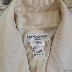 Puede incluir: Blazer Yves Saint Laurent color crema. El blazer presenta una tela texturizada y acanalada y un cuello con muescas. La etiqueta interior dice "MADE IN FRANCE" e incluye instrucciones de lavado. La talla es USA 12.