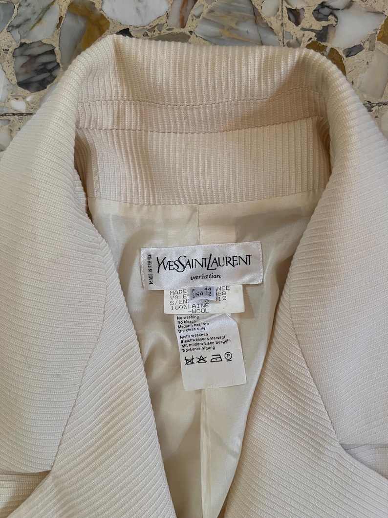 Puede incluir: Blazer Yves Saint Laurent color crema. El blazer presenta una tela texturizada y acanalada y un cuello con muescas. La etiqueta interior dice "MADE IN FRANCE" e incluye instrucciones de lavado. La talla es USA 12.