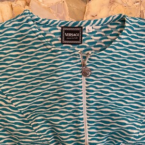 Op de afbeelding: Een teal en wit gestreept ritsjack met een Versace Jeans Couture label en een zilveren rits met het Versace logo.