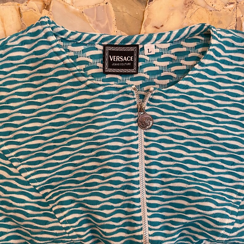 Op de afbeelding: Een teal en wit gestreept ritsjack met een Versace Jeans Couture label en een zilveren rits met het Versace logo.