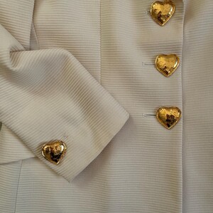 Puede incluir: Primer plano de una blazer beige claro con botones dorados en forma de coraz&oacute;n. La blazer tiene una tela texturizada y un dise&ntilde;o cl&aacute;sico. Los botones dorados brillantes a&ntilde;aden un toque de elegancia a la prenda.