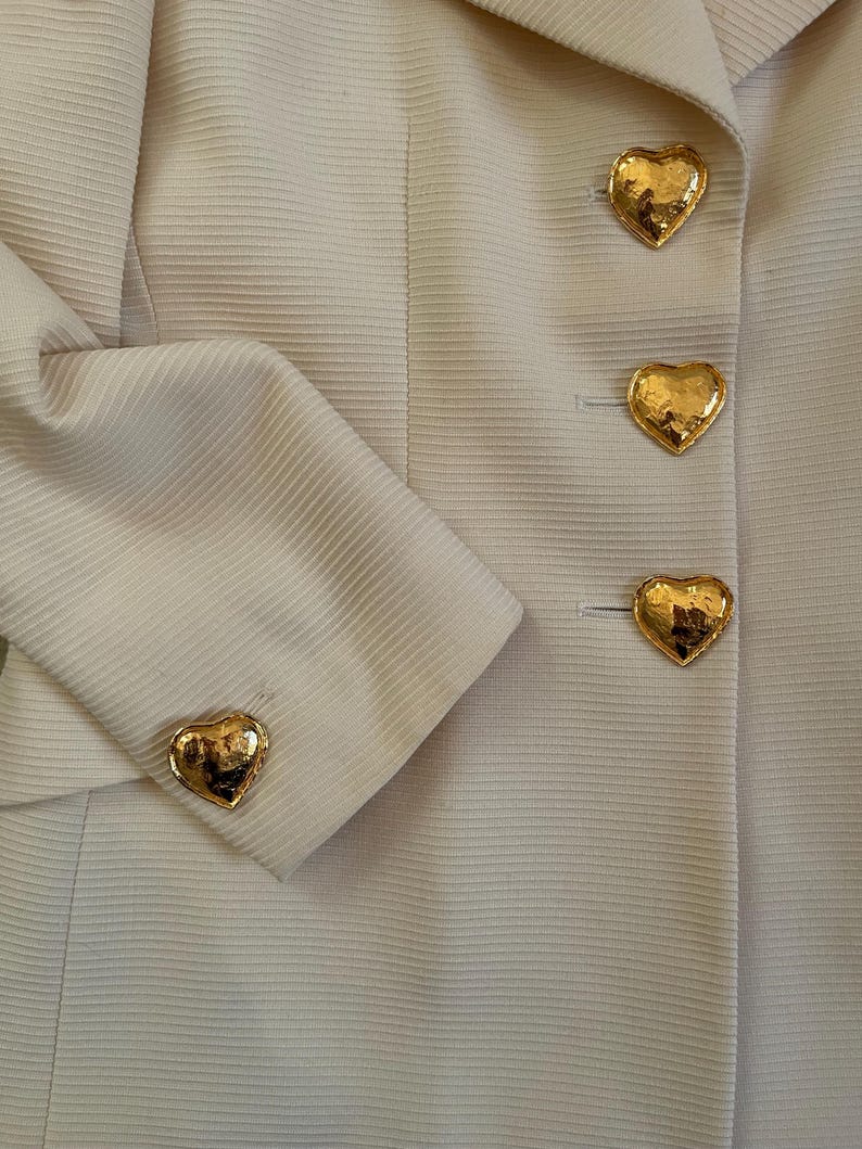 Puede incluir: Primer plano de una blazer beige claro con botones dorados en forma de coraz&oacute;n. La blazer tiene una tela texturizada y un dise&ntilde;o cl&aacute;sico. Los botones dorados brillantes a&ntilde;aden un toque de elegancia a la prenda.