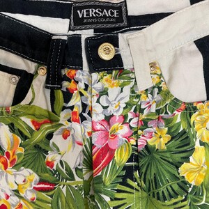 May include: Versace Jeans Couture pants featuring a black and white striped waistband with a gold-tone button closure. The lower portion displays a vibrant tropical print with green palm leaves, yellow hibiscus, and pink and white orchid flowers.