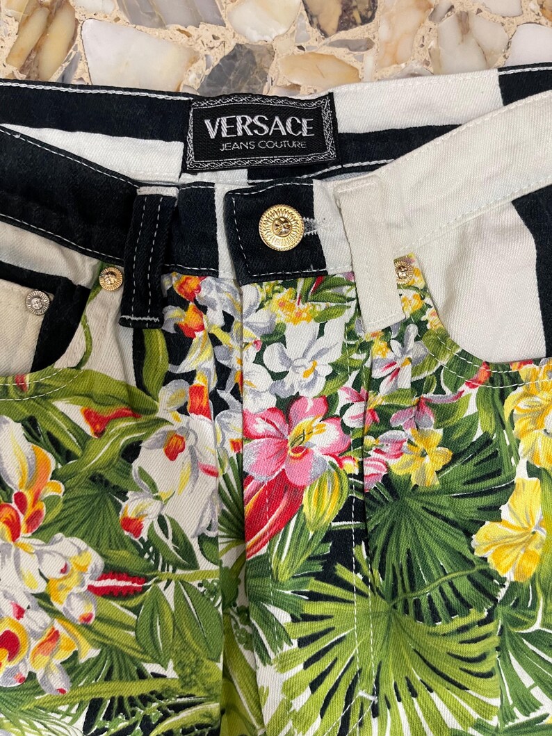May include: Versace Jeans Couture pants featuring a black and white striped waistband with a gold-tone button closure. The lower portion displays a vibrant tropical print with green palm leaves, yellow hibiscus, and pink and white orchid flowers.
