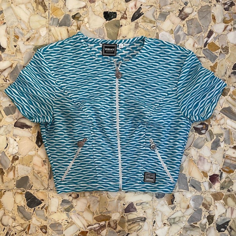Op de afbeelding: Een kort, blauw-wit gestreept, met rits te sluiten topje met het woord "VERSACE" op de rits.
