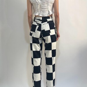 May include: A white sleeveless top with a ruffled neckline and a lace-up back closure paired with high-waisted pants featuring a bold black and white checkered pattern. The pants include two back pockets and a standard button and zipper fly closure.