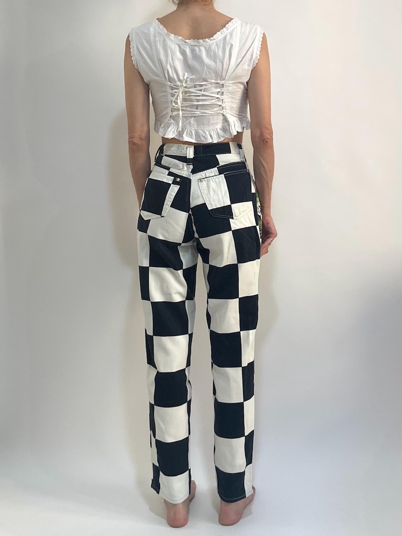 May include: A white sleeveless top with a ruffled neckline and a lace-up back closure paired with high-waisted pants featuring a bold black and white checkered pattern. The pants include two back pockets and a standard button and zipper fly closure.