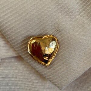 Puede incluir: Un bot&oacute;n dorado en forma de coraz&oacute;n con una superficie texturizada. El bot&oacute;n est&aacute; colocado sobre una tela beige claro con un sutil patr&oacute;n acanalado. El bot&oacute;n est&aacute; sujeto a la tela.