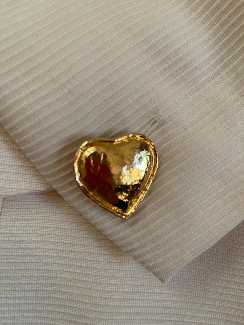 Puede incluir: Un bot&oacute;n dorado en forma de coraz&oacute;n con una superficie texturizada. El bot&oacute;n est&aacute; colocado sobre una tela beige claro con un sutil patr&oacute;n acanalado. El bot&oacute;n est&aacute; sujeto a la tela.