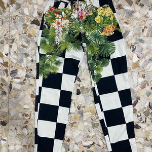 May include: High-waisted jeans featuring a black and white checkered pattern across the legs. The upper front section displays a vibrant botanical print with green palm leaves, yellow hibiscus, and white orchid flowers. A Versace label is visible.