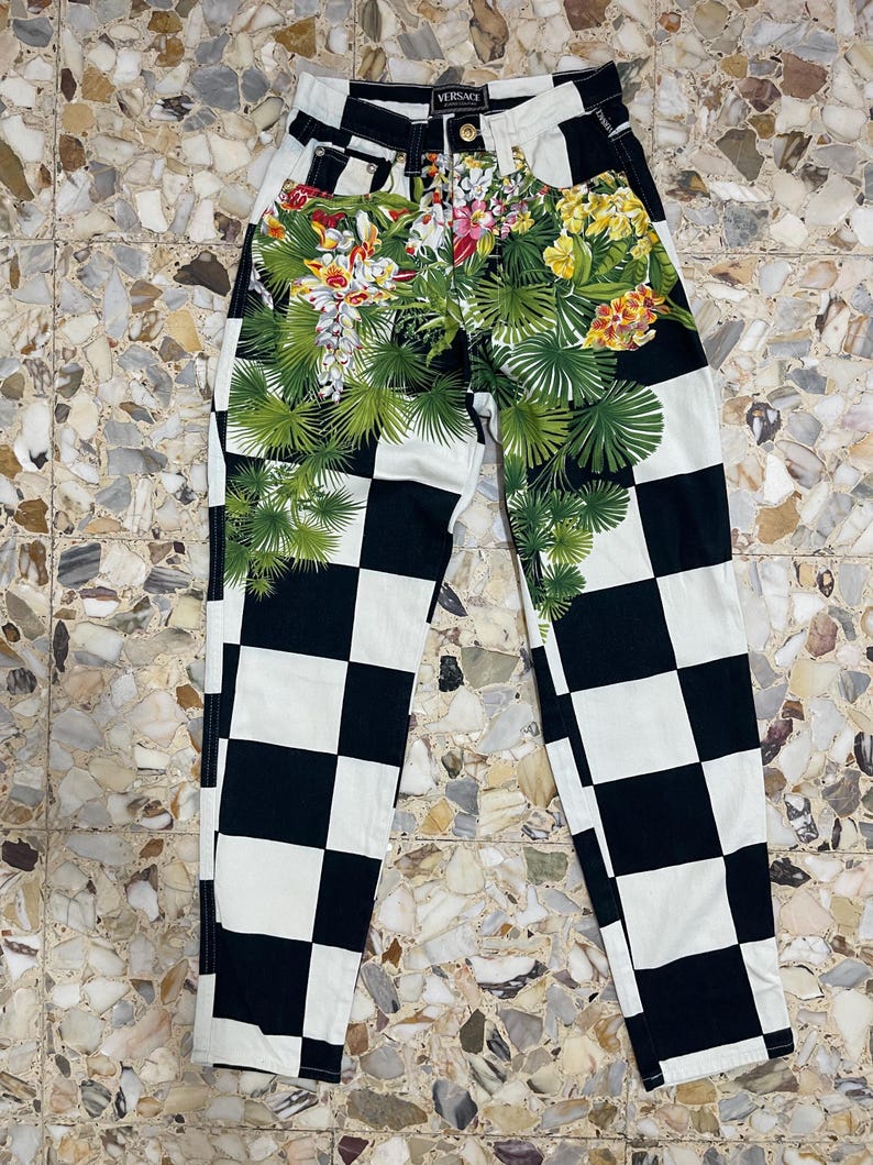 May include: High-waisted jeans featuring a black and white checkered pattern across the legs. The upper front section displays a vibrant botanical print with green palm leaves, yellow hibiscus, and white orchid flowers. A Versace label is visible.