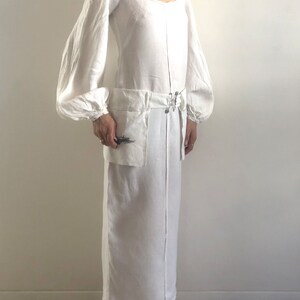 May include: A white linen dress with long, puffed sleeves and a scoop neck. The dress has a cinched waist with a belt and a large pocket on the front.