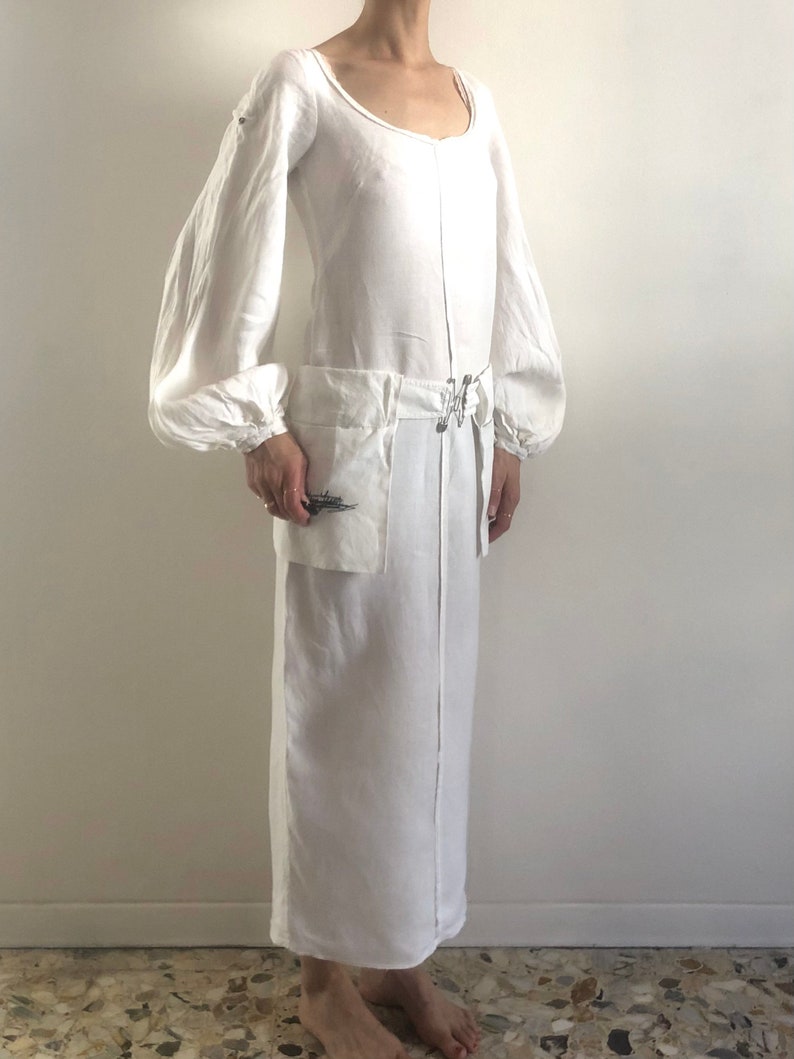May include: A white linen dress with long, puffed sleeves and a scoop neck. The dress has a cinched waist with a belt and a large pocket on the front.