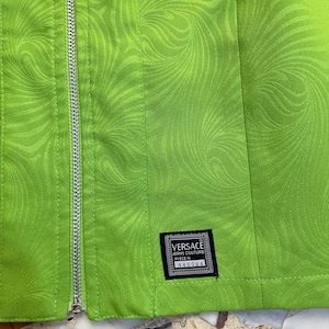 Może przedstawiać: Zielona sp&oacute;dnica z wirującym wzorem i srebrnym zamkiem błyskawicznym. Sp&oacute;dnica ma czarną metkę z tekstem "VERSACE JEANS COUTURE PIECE N. 440064".