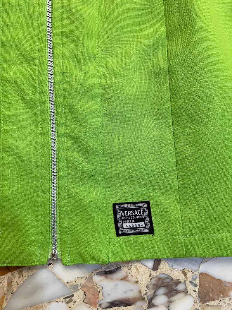 Może przedstawiać: Zielona sp&oacute;dnica z wirującym wzorem i srebrnym zamkiem błyskawicznym. Sp&oacute;dnica ma czarną metkę z tekstem "VERSACE JEANS COUTURE PIECE N. 440064".