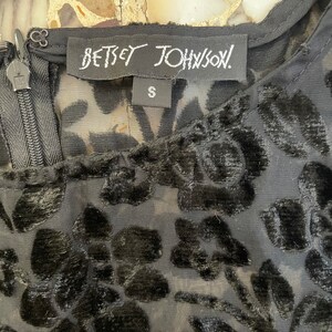 Może przedstawiać: Czarny, przezroczysty top z aksamitnym wzorem kwiatowym i metką Betsey Johnson. Metka zawiera napis "Betsey Johnson" i "S" oznaczający rozmiar.