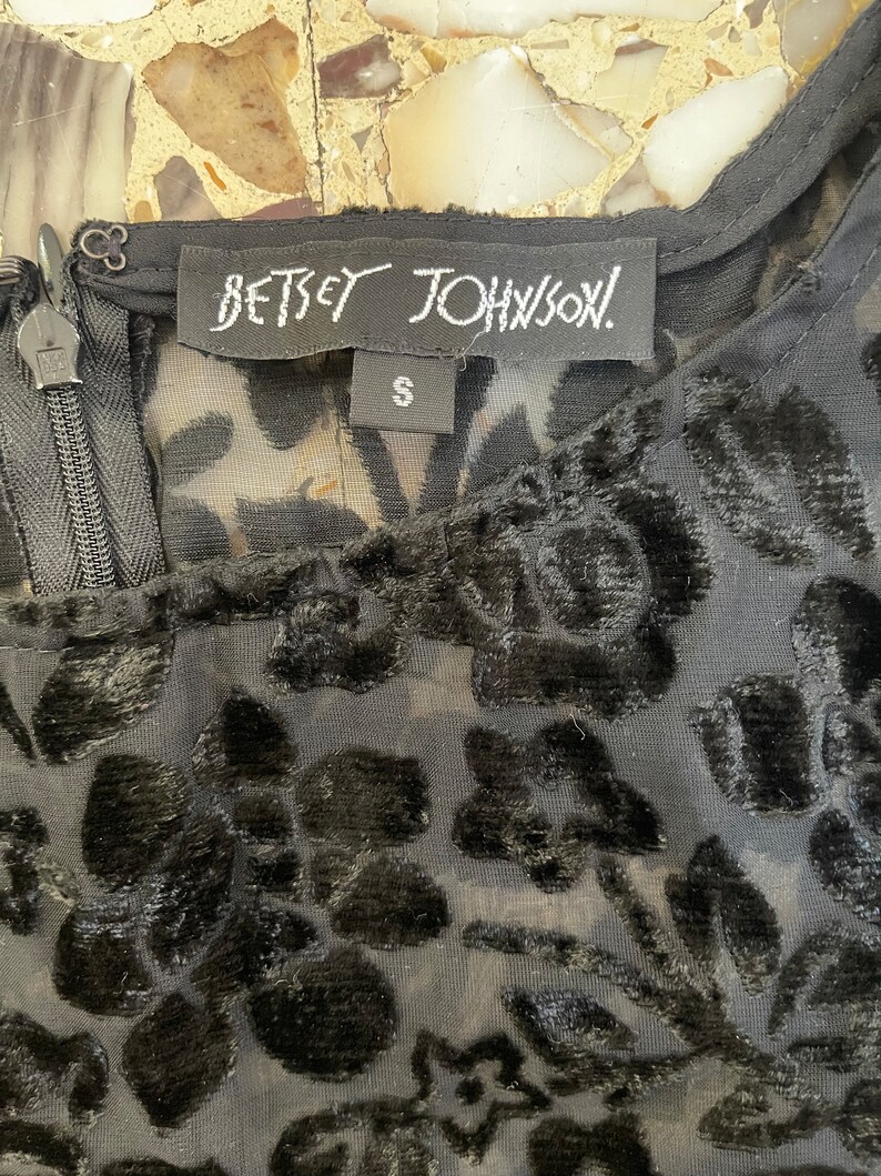Może przedstawiać: Czarny, przezroczysty top z aksamitnym wzorem kwiatowym i metką Betsey Johnson. Metka zawiera napis "Betsey Johnson" i "S" oznaczający rozmiar.