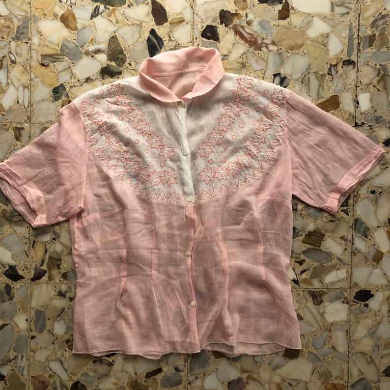 Pu&ograve; includere: Una camicia rosa a maniche corte con bottoni e ricami bianchi. Il ricamo &egrave; un motivo floreale con un bordo a festoni.